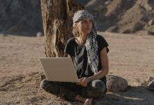 Caucasian woman sitting barefoot with laptop under tree in arid landscape.