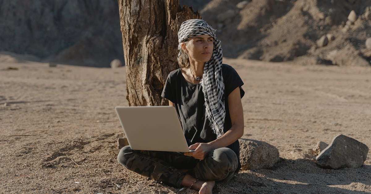 Como Criar uma Rotina Produtiva Enquanto Viaja pelo Mundo - Web Nômade Digital Caucasian woman sitting barefoot with laptop under tree in arid landscape.