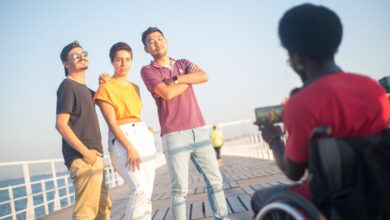 Como Viajar Sustentavelmente: Dicas para Nômades Digitais - Web Nômade Digital A joyful group of friends posing on a boardwalk, captured by a photographer in a wheelchair on a sunny day.