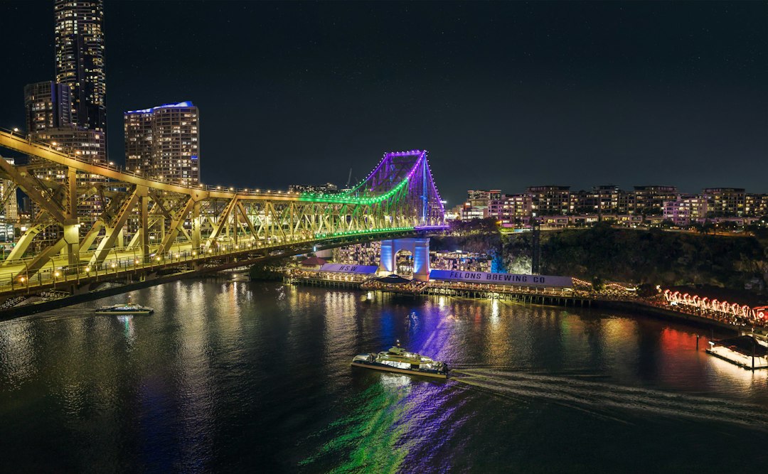 Como Gerenciar seu Orçamento de Viagem com Apps Práticos - Web Nômade Digital The Story Bridge illuminated at night above the Brisbane River with a ferry